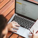 Over head shot of someone using laptop on top of a wooden table whilst looking at phone screen